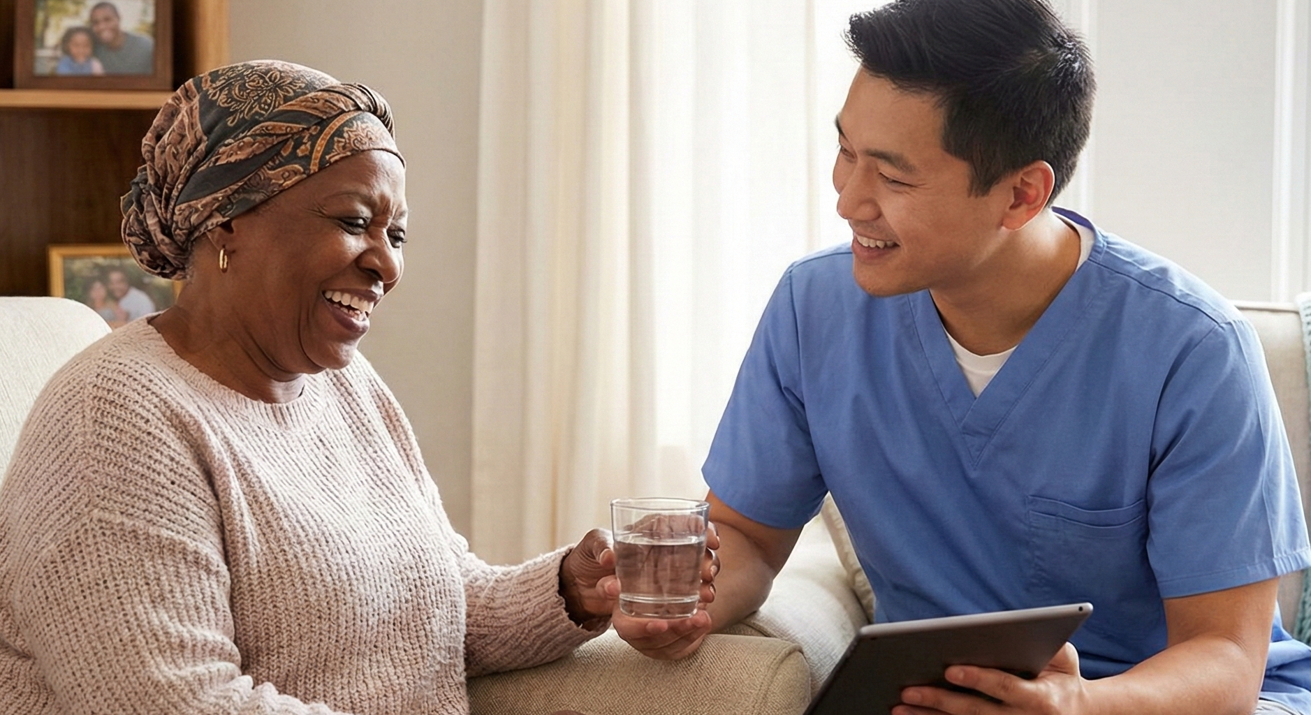 Smiling woman in headscarf at home accepts a glass of water from a clinician in scrubs holding a tablet during a supportive care visit.